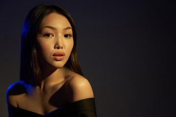 Fototapeta premium Portrait of a young beautiful Asian girl looking directly at the camera. The woman's hair and shoulders in colorful bright UV blue lights. On black background. 