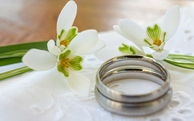 Frühlingsgefühle - Silberne Eheringe, in deren Innenseite Text geschrieben werden kann, liegen auf weißer Spitze neben Schneeglöckchen auf einem Holztisch