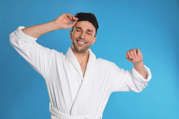 Naklejka premium Happy young man in bathrobe and eye sleeping mask on light blue background