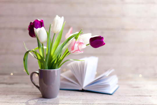 Spring flowers and book at wooden background. Tulips in gray cup. Bouquet in vase. Pink, white, lilac and purple blooming flora. Cozy still life. Copy space.