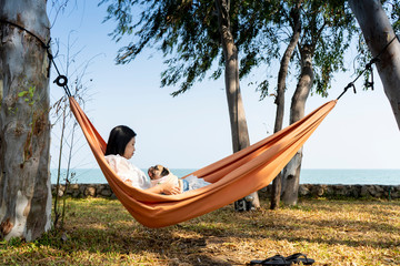 Funny cute pug dog smiling and tongue sticking out while on a trip with her owner, joyful young family, woman lying rest in comfortable hammock on sea beach. Happy times concept.