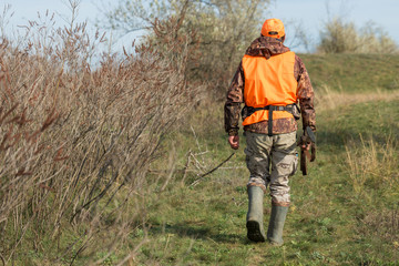 A man with a gun in his hands and an orange vest on a pheasant hunt in a wooded area in cloudy weather. Hunter with dogs in search of game.
