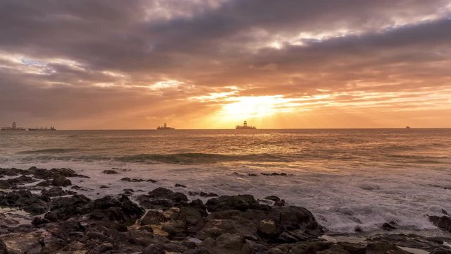 Time-lapse of sunrise from a stony shore to boats moored on the horizon with the rising sun and pesky shining through the clouds to the ocean with moving clouds from the Grand Canary Island.