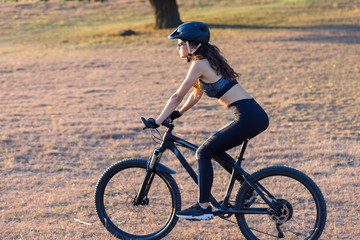 Fototapeta premium Girl on a mountain bike on offroad, beautiful portrait of a cyclist at sunset, Fitness girl rides a modern carbon fiber mountain bike in sportswear. Close-up portrait of a girl in a helmet and glasses