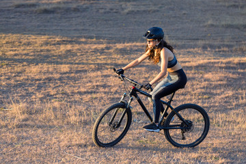 Fototapeta na wymiar Girl on a mountain bike on offroad, beautiful portrait of a cyclist at sunset, Fitness girl rides a modern carbon fiber mountain bike in sportswear. Close-up portrait of a girl in a helmet and glasses