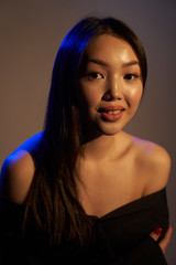  Portrait of a young beautiful Asian girl looking directly at the camera. The woman's hair and shoulders in colorful bright UV blue lights. On black background.  