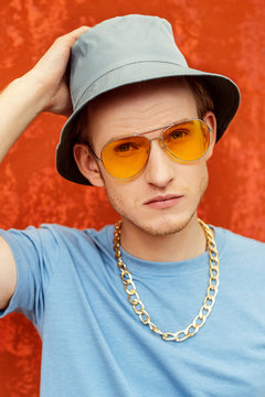 Close up portrait of young fashionable handsome man wearing orange color sunglasses, blue bucket hat, t-shirt, golden chain, posing against colorful background