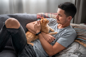 un hombre joven recostado en un sofa abraza a un gato atigrado. Ambos se miran uno a otro con amor - obrazy, fototapety, plakaty