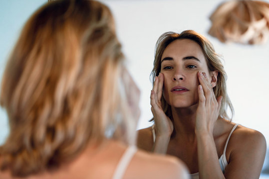 Close-up of young woman touching her cheekbones while looking in mirror in bathroom