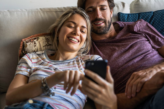 Smiling couple using smartphone while relaxing on sofa at home