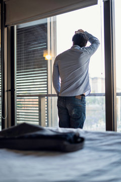 Rear view of thoughtful young man looking through window with blazer on bed in foreground