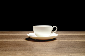 White glossy ceramic cup on saucer placed on wooden table with black background