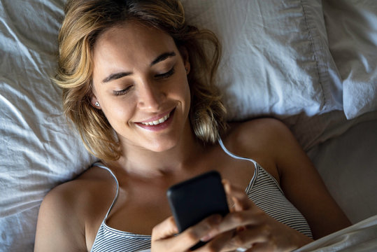 Close-up of smiling young woman using smartphone while lying on bed in bedroom
