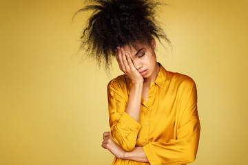 Tired girl covers face with palm, feels overworked and fatigue. Photo of african american girl on yellow background