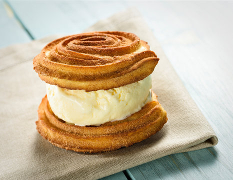 Churro Ice Cream Sandwich, Made With Homemade Churros And Vanilla Ice Cream. A Delicious Dessert Served On A Bege Napkin On A Blue Wooden Table.