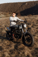 Fototapeta na wymiar Vertical view. Handsome male in eyeglasses and gloves motorcyclist travels on motorbike, treveler in desert, looking at a side in sunset time.