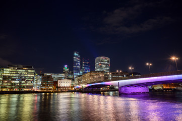City of London, UK with view over the River Thames and London Bridge at night. Wide cityscape with skyscrapers, lights, and dark sky. London 2020.