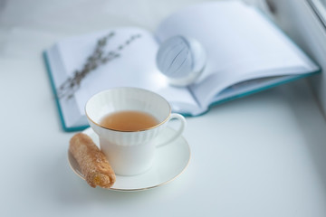 A white Cup with tea on a saucer is a cookie, on an open book is a glass ball on a white background. Selective focus.
