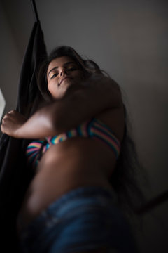 Low angle portrait of young and beautiful dark skinned Indian Bengali woman in colorful lingerie/bikini and hot pants standing in front of window and looking outside. Boudoir photography.
