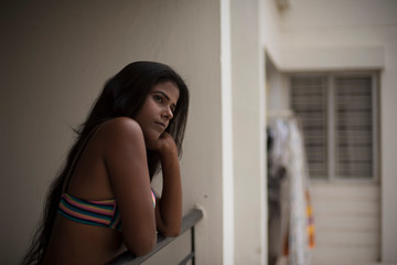 Fototapeta na wymiar Portrait of an young and beautiful dark skinned Indian Bengali woman in colorful lingerie/bikini and hot pants posing in casual mood on a balcony in white urban background. Boudoir photography.