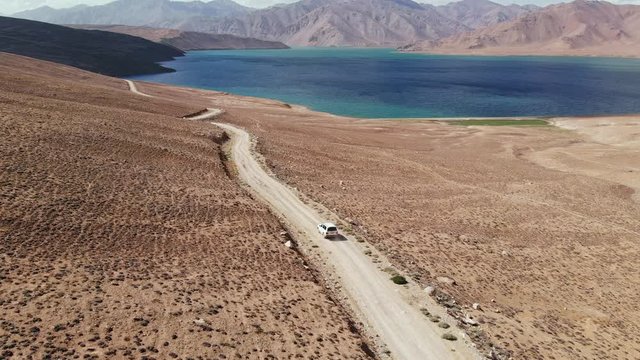 Aerial over off road 4x4 car driving along gravel trail path toward lake in arid mountains.Pamir Highway silk road travelling adventure in Tajikistan,Asia.Khorugh Bulunkul lake. 4k drone flight video