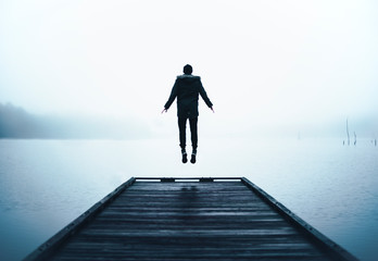 Man Jumping in Air Levitating at End of Wooden Dock Over Big Lake with Thick Layer of Fog and Clouds at Sunrise on Cold Brisk Wet Morning   - obrazy, fototapety, plakaty