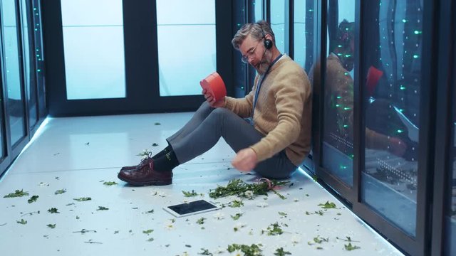 Frustrated IT technician crying of one-sided love holding a present box sitting on floor covered with flowers worrying in server room. Sad Valentine's Day in data center.
