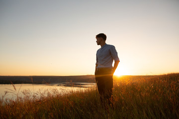 Man standing and enjoying the beautiful sunset over a wide river valley. Young man in  blue shirt...