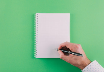 top view of hand holding ballpoint pen against spiral notepad on green background, taking notes concept