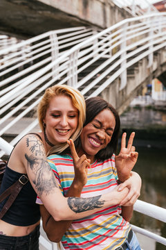 two girl friends in a embrace, one with tattooed arms hugging a brunette woman doing peace sing.