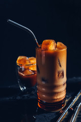 Ice coffee from ice cubes with milk in a tall glass, dark mood, metal zero waste straws for cocktails, a jar of coffee beans, black background