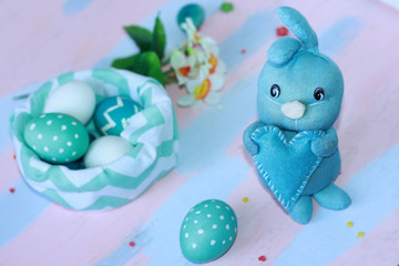 Easter light blue rabbit stands with a blue heart in its paws near the basket with blue-white eggs. Handmade. Greeting card for happy easter.