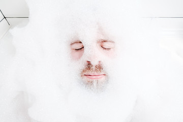 Male face in a lush white foam, only eyes and mouth are visible, close-up. Man relaxes while lying in a bathtub, with soapy water.