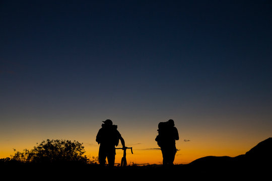 two men with backpacks and bicycle walking into the sunset on mountain