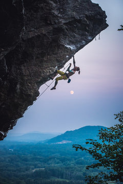 Man clipping quickdraw on overhanging sport climbing route with moon.