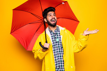 Young handsome bearded indian man in yellow raincoat with red umbrella cover from rain over orange background