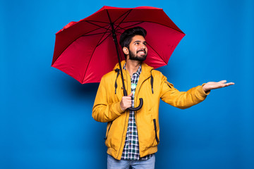 Young handsome bearded indian man in yellow raincoat with red umbrella cover from rain isolated...