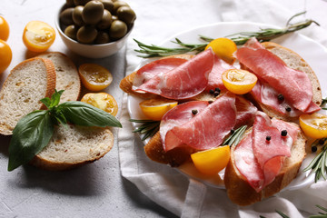 The concept of Italian cuisine. Coppa - Italian dried sausage made from pork neck. Meat snacks. Baguette, olives, cherry tomatoes, rosemary, basil on a light white background.  copy space