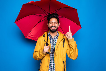 Young handsome bearded indian man in yellow raincoat with red umbrella cover from rain have idea isolated over blue background