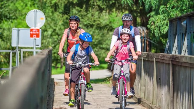 Family cycles over a wooden bridge. Sun shines on a summer day. Slowmotion. Slovenj Gradec