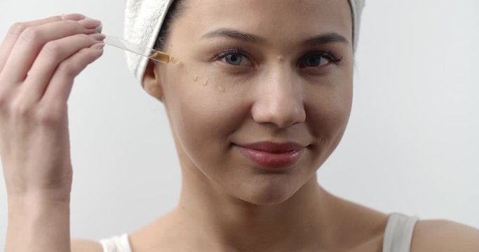 Portrait of attractive young lady with towel on head applying essential oil under eyes from pipette. Isolated over white background. Concept of facial skin care.