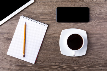 Obraz na płótnie Canvas Smartphone and tablet PC with black screens on a wooden background with cup of black coffee. An opened notebook with pencil on it lie next to them.
