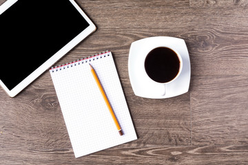 Tablet PC with black screen on a wooden background with cup of black coffee. An opened notebook with a pencil on it lie next to them.