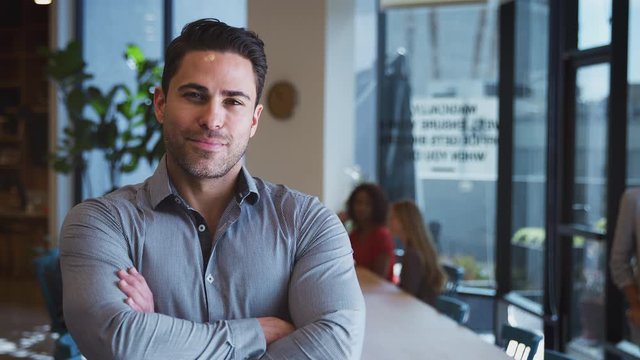 Mature businessman in modern open plan office work space smiling into camera with colleagues in background - shot in slow motion