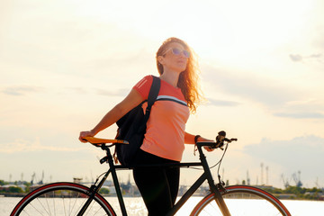 After cycling young red-haired woman stands with bicycle on pier of city seaport against background of ships and cranes at sunset in orange sun, looks into horizon. Lifestyle