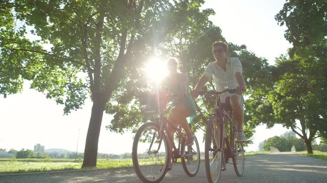 SLOW MOTION, LENS FLARE, LOW ANGLE: Sun rays shine on happy tourist couple riding their bicycles around a picturesque park. Cute couple enjoys a sunny summer day by going for a relaxing bike ride.
