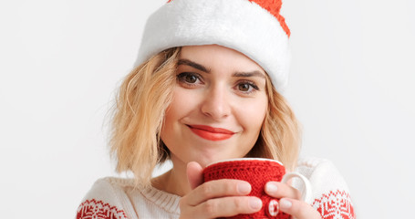 Young blond woman in Christmas red cap hat holds and blows mug of hot cocoa, drinks drink, smiles, warms her hands on white background