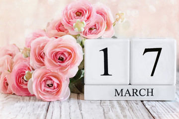 Fototapeta premium White wood calendar blocks with the date March 17th and pink ranunculus flowers over a wooden table. Selective focus with blurred background. St Patrick Day.
