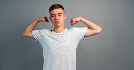 Portrait of confident sports teenager boy showing her biceps triceps grey background raising arms up muscle mass. Athlete shows fitness camera, is happy to lower hands down. Sports concept