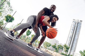 Outdoors Activity. African couple on basketball court girl dribbling while guy hugging blocking her...